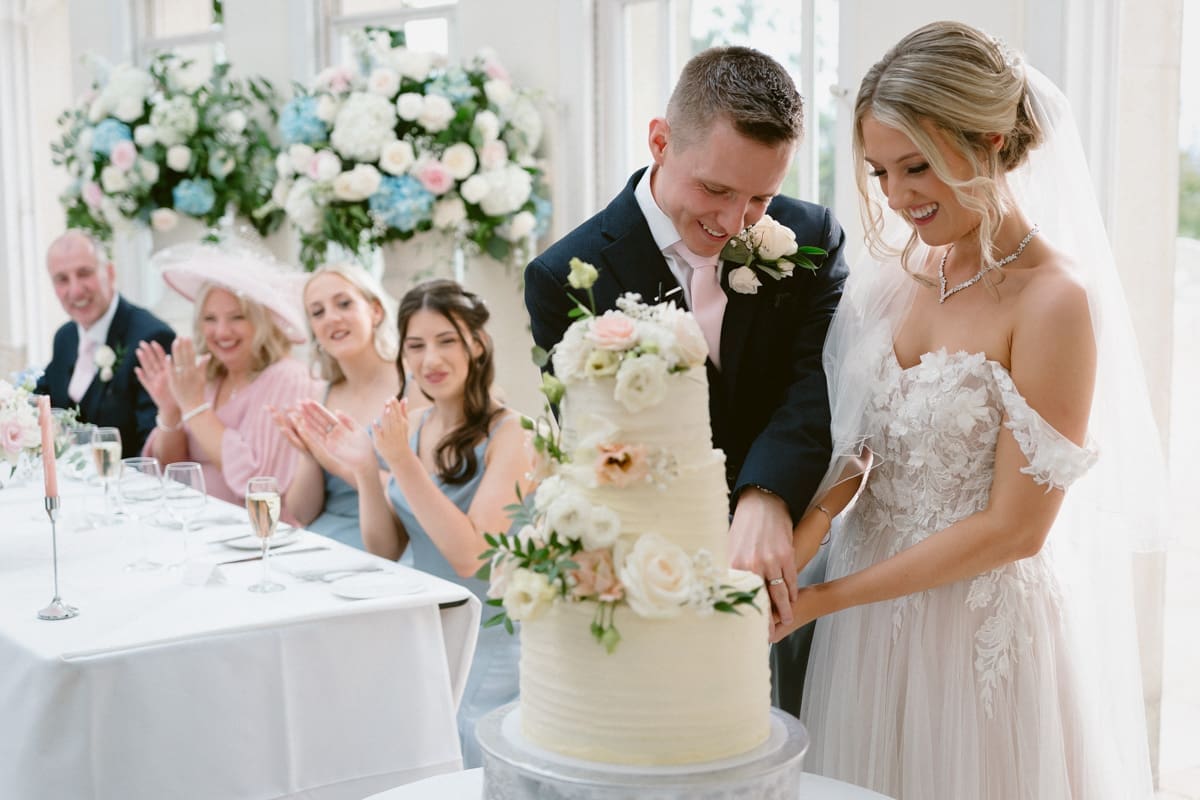newly married couple cut their wedding cake while family look on.